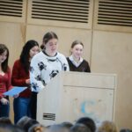 Young woman speaks at a wooden podium in a beige auditorium, with peers standing behind her and an audience in the foreground.