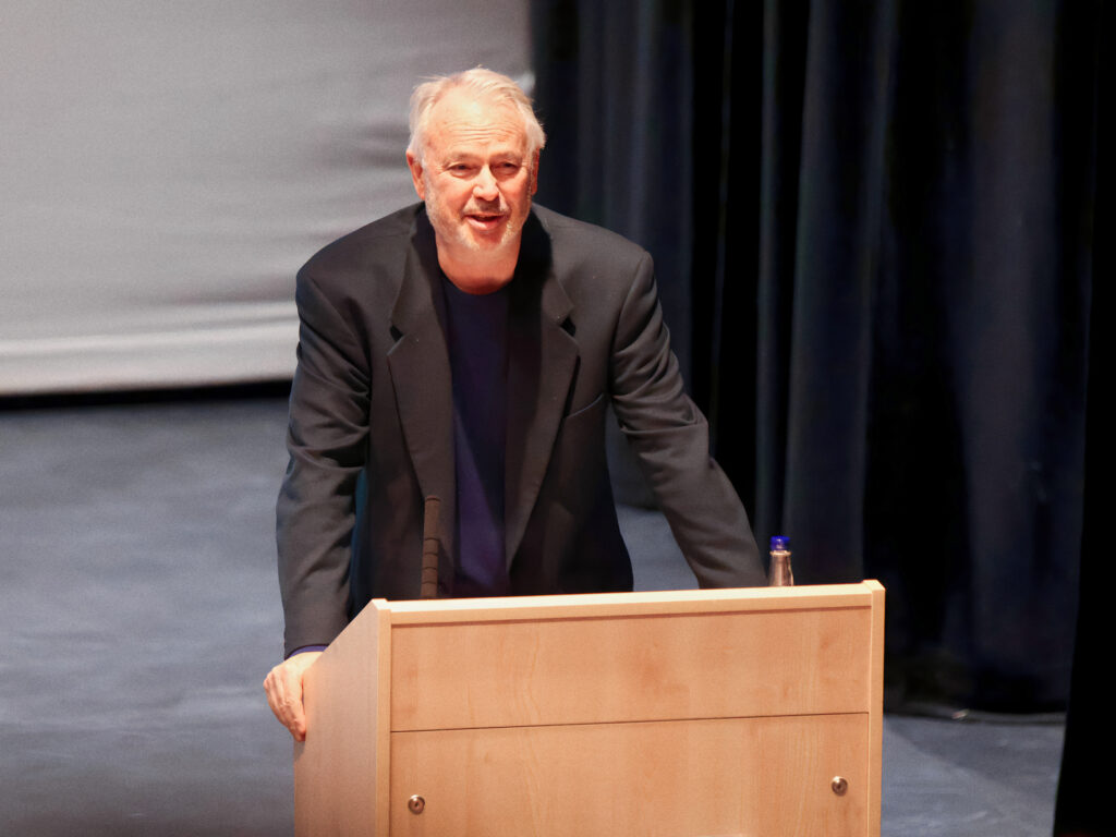 Older man in a dark blazer leaning on a wooden podium, speaking on a stage with black curtains in the background.
