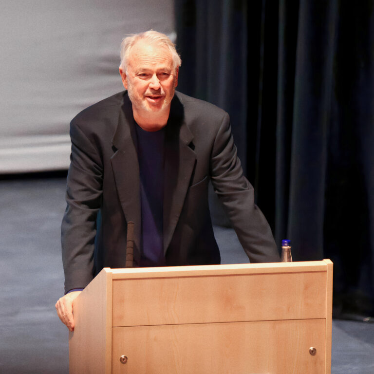 Older man in a dark blazer leaning on a wooden podium, speaking on a stage with black curtains in the background.
