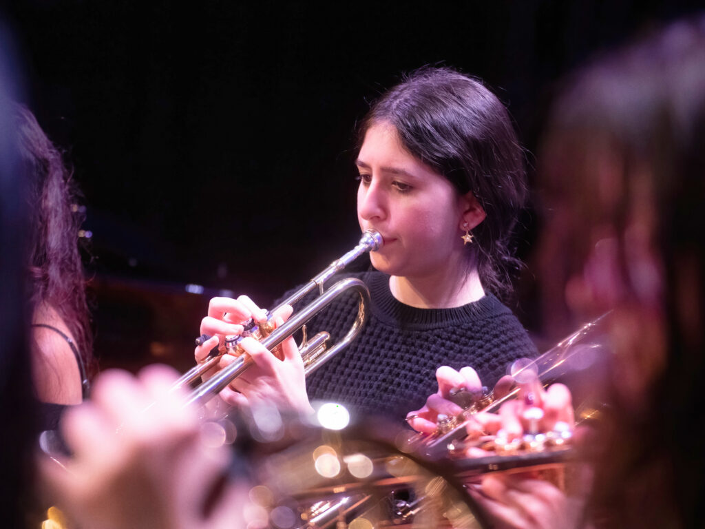 Woman playing a silver trumpet, focused on notes, wearing a black knit sweater; other musicians blur in the foreground.