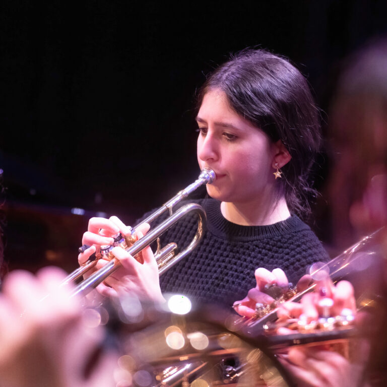 Woman playing a silver trumpet, focused on notes, wearing a black knit sweater; other musicians blur in the foreground.