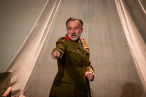 Actor in a vintage military uniform on stage, pointing emphatically as he delivers a dramatic line.