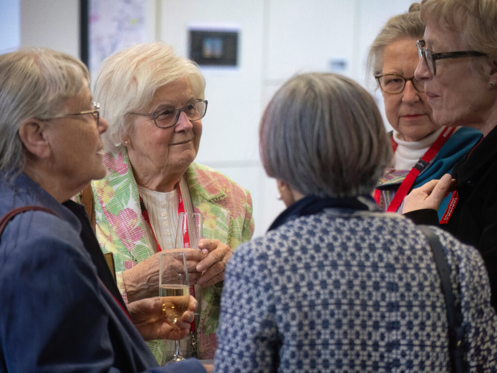 Group of elderly women in a social gathering, chatting closely and holding drinks in a bright room.