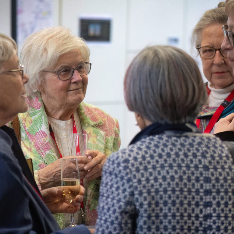 Group of elderly women in a social gathering, chatting closely and holding drinks in a bright room.