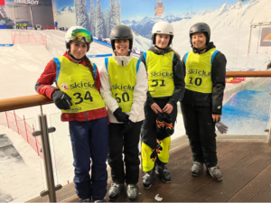 Four young skiers in helmets and neon bibs posing on a ski slope area, bib numbers 34, 50, 51 and 20.