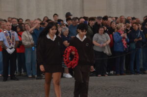 Two young students in brown uniforms carry red wreaths as they walk past a large crowd at a ceremony.