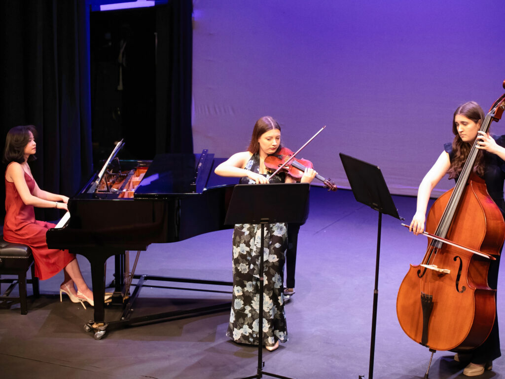Three female musicians performing on a stage: a pianist in a red dress on the left, a violinist in a floral dress center, and a double bassist on the right under purple lighting.