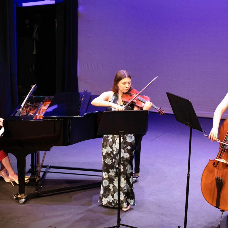 Three female musicians performing on a stage: a pianist in a red dress on the left, a violinist in a floral dress center, and a double bassist on the right under purple lighting.
