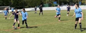 Youth girls' soccer match: two players in blue and navy vie for the ball on a sunny grassy field, with teammates in the background.