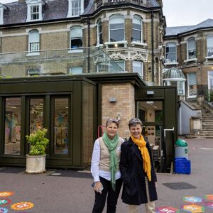 Two smiling women stand outside a brick historic building, with colorful chalk-number shapes drawn on the pavement around them.