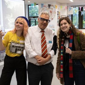 Three adults posing together at a social event, each holding a drink, indoors with colorful bunting in the background with a projector screen nearby