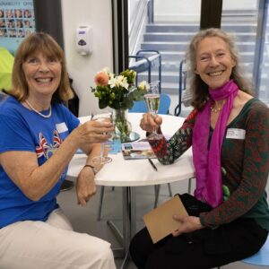 Two smiling women toast with wine glasses at a round table, a small flower arrangement between them in a bright indoor setting.