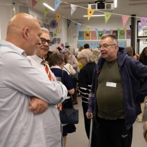 People converse at a festive indoor gathering with colorful bunting, a projector overhead, and tables set with drinks in the background.
