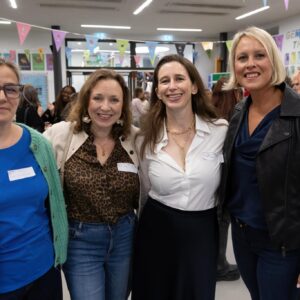Four women stand together smiling at the camera in an indoor event space with colorful bunting banners overhead.