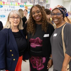 Three women stand together, smiling at a community or school event indoors.