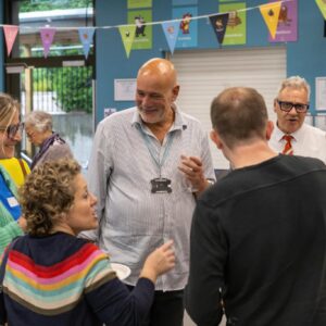 Group of adults chatting and laughing at an indoor community event with colorful bunting overhead.