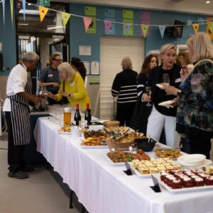 Guests mingle around a long buffet table covered with a white cloth, with assorted appetizers and desserts and wine glasses on display in a bright room with bunting overhead.