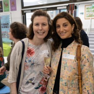 Two women smile for a photo at an indoor event, each holding a wine glass and wearing name badges with floral outfits.