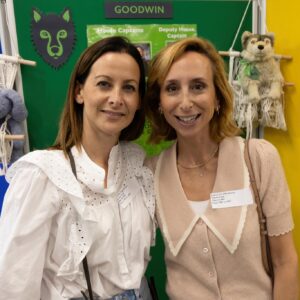 Two women smile for a photo at a colorful exhibition booth with banners and a stuffed lion in the background, wearing name badges on light clothing.