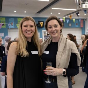 Two smiling women posing for a photo at an indoor social event, each wearing name tags and holding drinks behind them a bunting-decorated room.