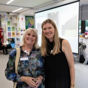 Two smiling women pose for a photo at an indoor event, with colorful posters and a projector screen in the background.