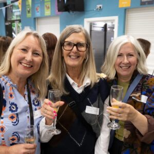 Three smiling women posing for a photo at an indoor celebration, each holding a wine glass in a toast edge-to-edge shot.