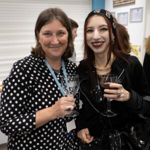 Two smiling women posing together, each holding a wine glass at an indoor event.