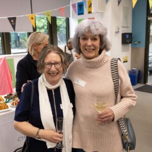 Two elderly women smile at a social event, each holding a wine glass, with colorful bunting and a food table in the background.