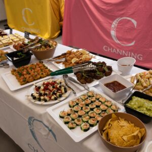 A long banquet table covered with a white cloth, loaded with bite-sized appetizers, skewers, wraps, dips, and a bottle of wine, with pink branding in the background.