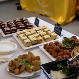 Assorted bite-sized desserts and appetizers arranged on a buffet table, including layered chocolate cakes and cream-topped squares with fruit nearby.