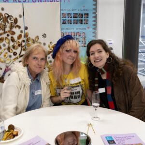 Three women sit together at a round table smiling for the camera at an indoor social event, with snacks and drinks on the table behind them.