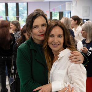 Two women hug and smile at a crowded indoor event, one in a green jacket and the other in a white shirt with a name badge visible on her chest