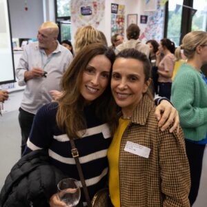 Two women smile for a selfie at a crowded social event, drinks in hand and other guests in the background.