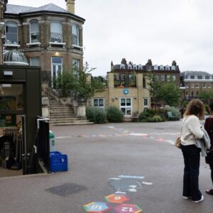 Two women stand in a courtyard with chalk hopscotch numbering on the ground and brick buildings in the background.