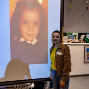 Smiling woman in a yellow top and plaid jacket stands beside a large projected portrait of a young girl with a white bow in her hair.