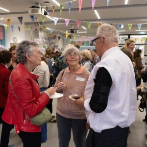 Group of people mingling at an indoor reception, holding plates and drinks under colorful pennant bunting in a decorated hall.