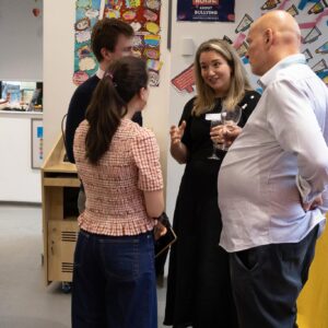 Group of adults chatting at a social or professional event near a yellow tablecloth display and flower arrangement in a classroom or conference space.