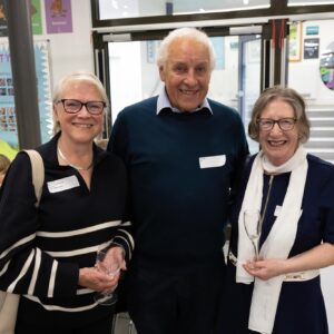 Three smiling adults with wine glasses pose together in a school hallway background and posters visible behind them. (Note: group consists of two women on either side and a man in the center.)