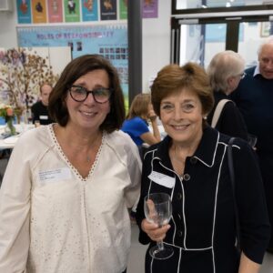 Two smiling women pose together at a reception, one in a white embroidered blouse and the other in a dark patterned jacket, each wearing a name badge and holding a wine glass.