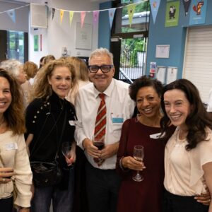 Group of adults at a social event, holding wine glasses, smiling for the camera in a room with colorful pennant banners overhead.