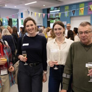 Four people pose for a photo at an indoor social event, each holding a glass of wine or juice, with colorful bunting overhead and other guests in the background.