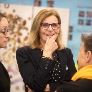 Center woman with glasses smiles and rests her finger on her chin during a group discussion in a bright indoor setting.