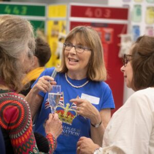Woman with glasses in a blue shirt holding a champagne flute, talking to a group at a social event for accessibility and clarity in context of gathering sizes.