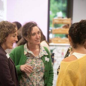 Three women stand in a small circle, smiling and chatting at an indoor social event.
