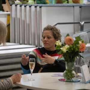 Woman in a colorful striped sweater speaks at a round table with a bouquet of pink and white flowers and a glass of champagne nearby.
