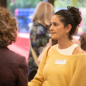 Profile view of a woman with dark curly hair in a bun, wearing a yellow sweater and a name badge at a social event.