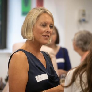 Side profile of a blonde woman in a navy dress speaking with others at an event; name badge visible on her chest.