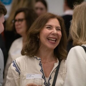 Smiling woman with wavy brown hair in a white and blue embroidered blouse, holding a glass at a social gathering.
