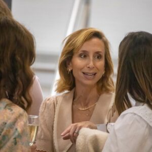 Three women in conversation at a social event, centered on a pale-haired woman in a beige cardigan smiling at two others.