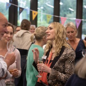People in a social gathering chatting and holding wine glasses under colorful bunting banners in a bright room.